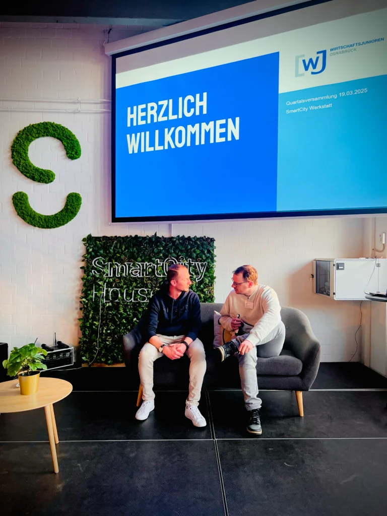 Zwei Personen sitzen auf einer Couch vor einem Bildschirm mit einer Präsentation der Wirtschaftsjunioren